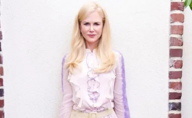 Blonde woman with straight hair in a pink and purple frilled blouse, standing against a white and brick background.