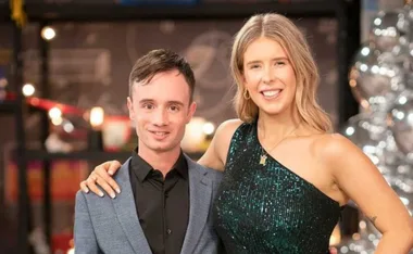 Two people embracing, wearing formal attire, smiling at the camera against a festive backdrop.