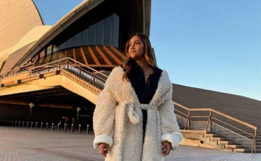 Woman in a fluffy coat stands in front of the Sydney Opera House under a clear sky.