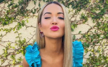 Woman with long blonde hair in a blue ruffled top winks and puckers lips, standing against a green vine wall.