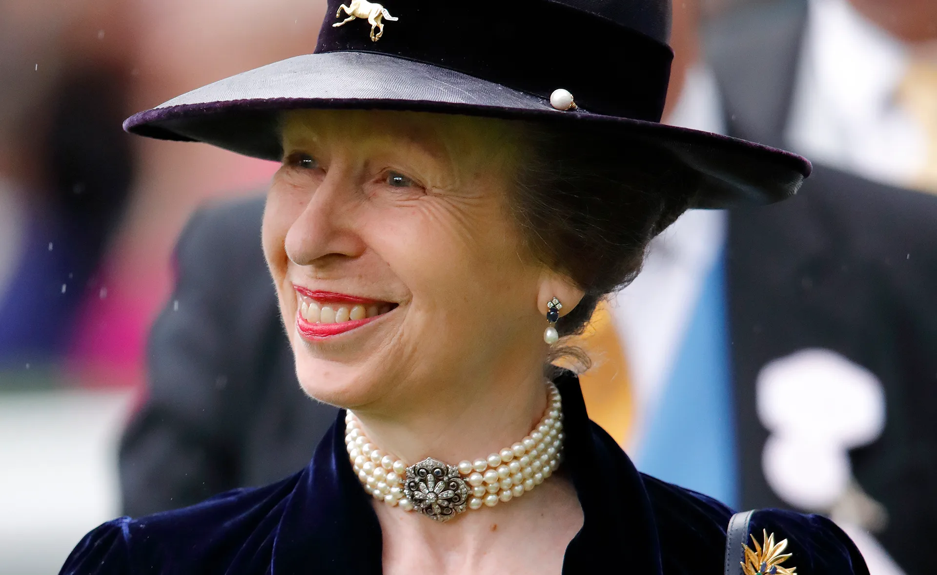 Smiling woman in a hat adorned with a horse pin, wearing a pearl choker and earrings against a blurred background.