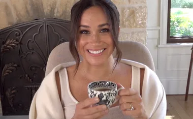 A woman smiling and holding a decorative mug in a cozy interior setting with a window and stone wall.