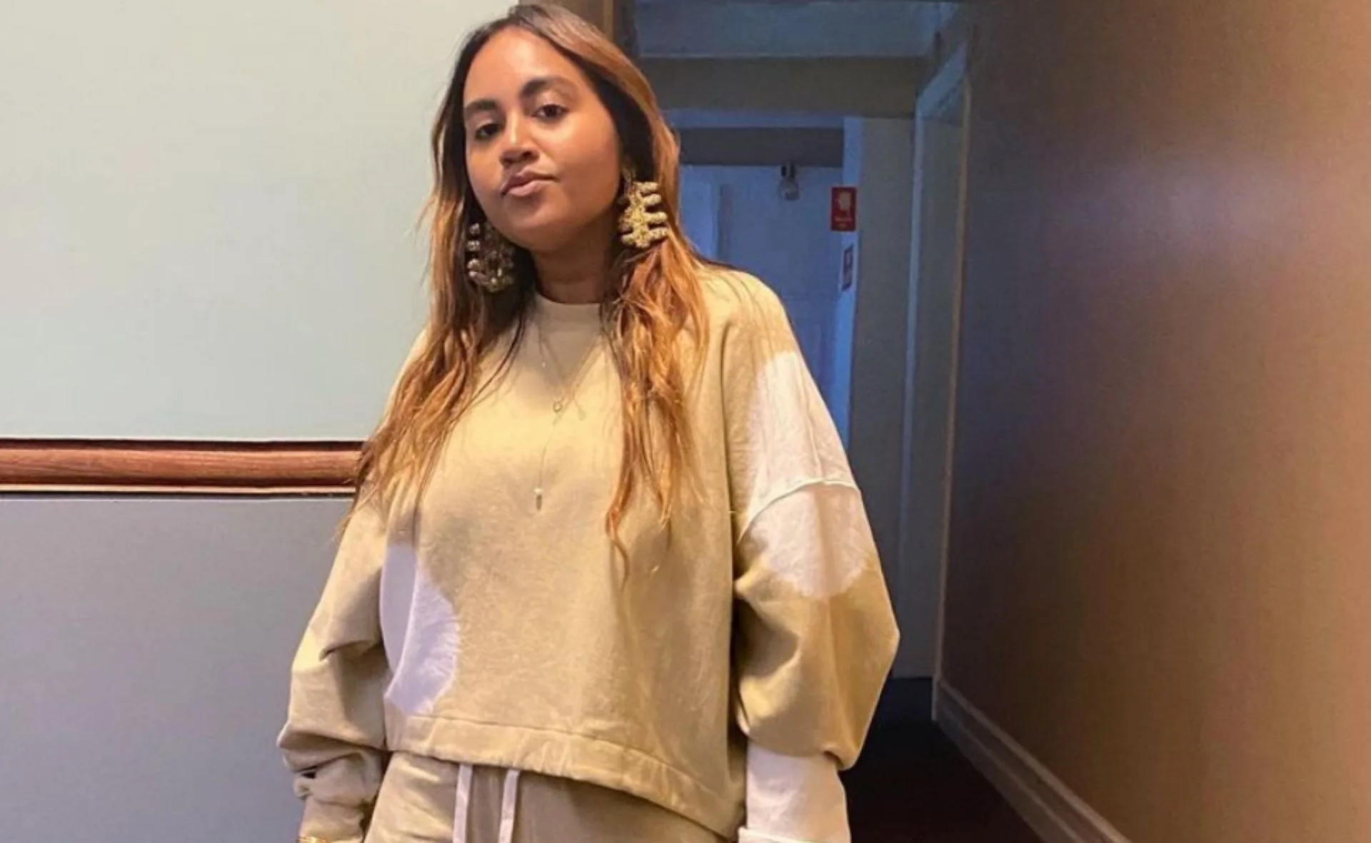 Woman in beige outfit and statement earrings stands in a hallway with soft lighting.