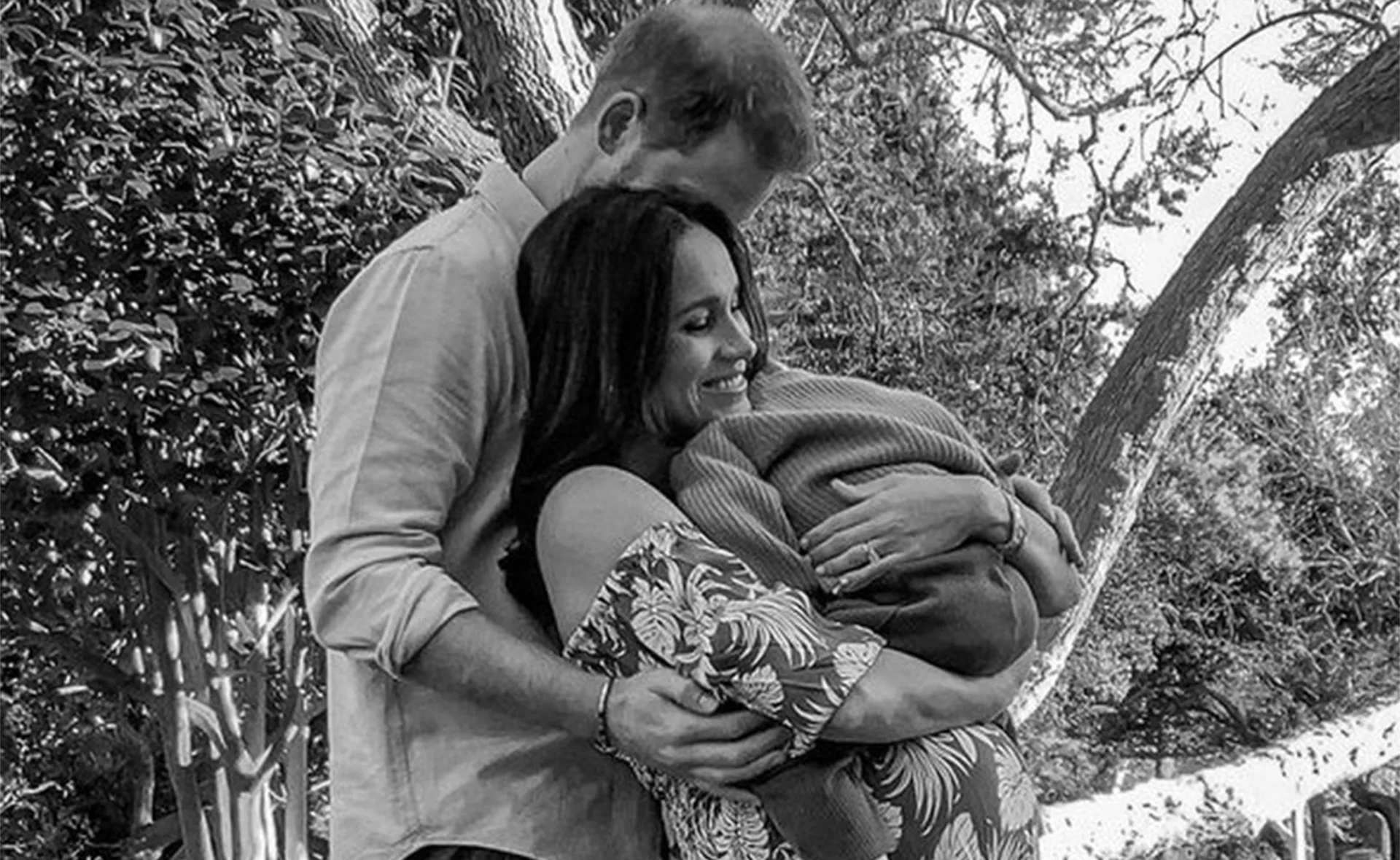 A couple embraces outdoors, the woman holds a baby wrapped in a blanket, surrounded by trees and greenery.
