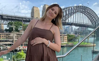 A woman in a brown dress poses smiling with the Sydney Harbour Bridge in the background.