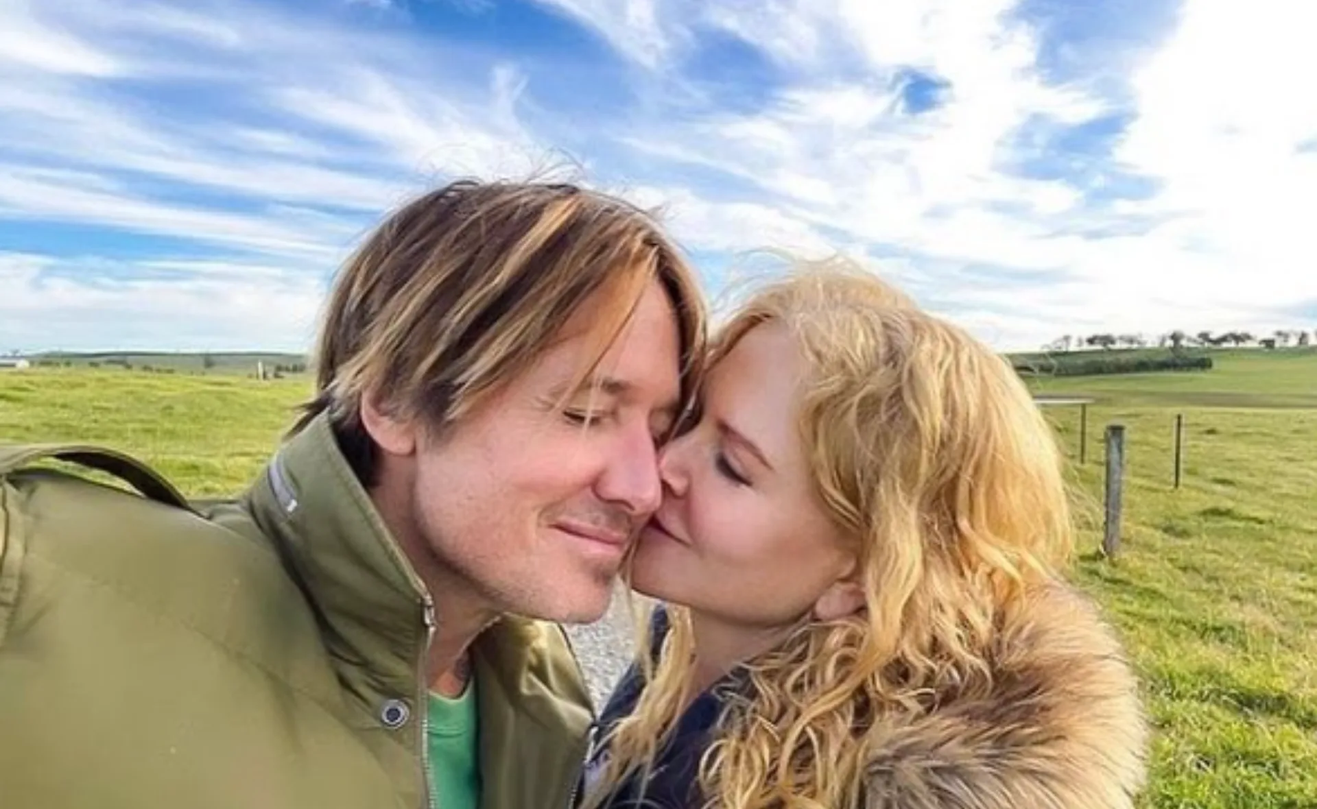 A couple embraces affectionately in a green field under a blue sky with clouds.