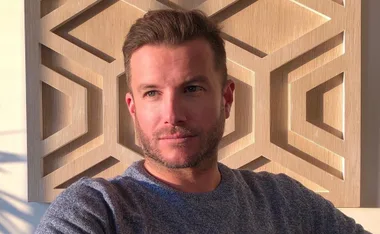 A man in a gray sweater sits in front of a geometric wooden wall, gazing to the side with sunlight highlighting his face.