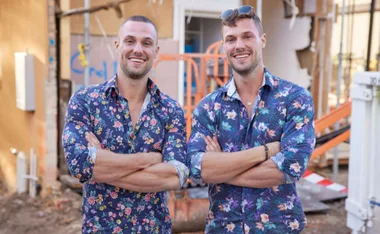Two smiling men in floral shirts stand with arms crossed at a construction site.