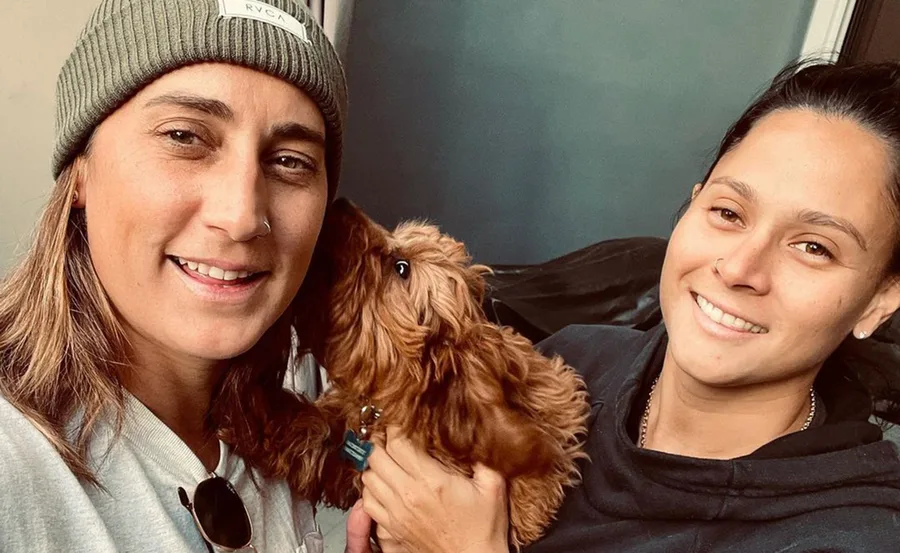 Two smiling individuals posing with a small brown dog indoors.