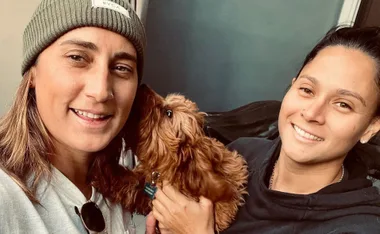 Two smiling individuals posing with a small brown dog indoors.