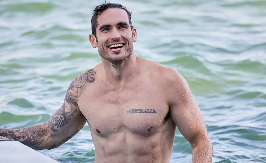 Man with tattoos smiling in water, visible text tattoo reads "AUSTRALIA."