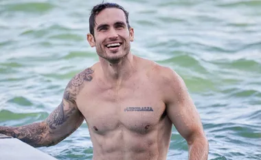 Man with tattoos smiling in water, visible text tattoo reads "AUSTRALIA."