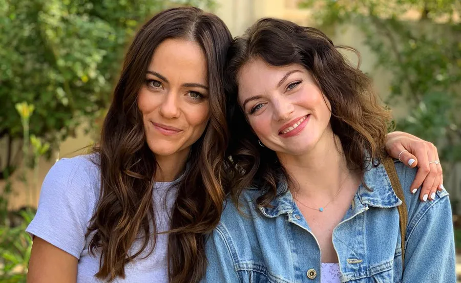 Two actresses smiling outdoors with greenery in the background.
