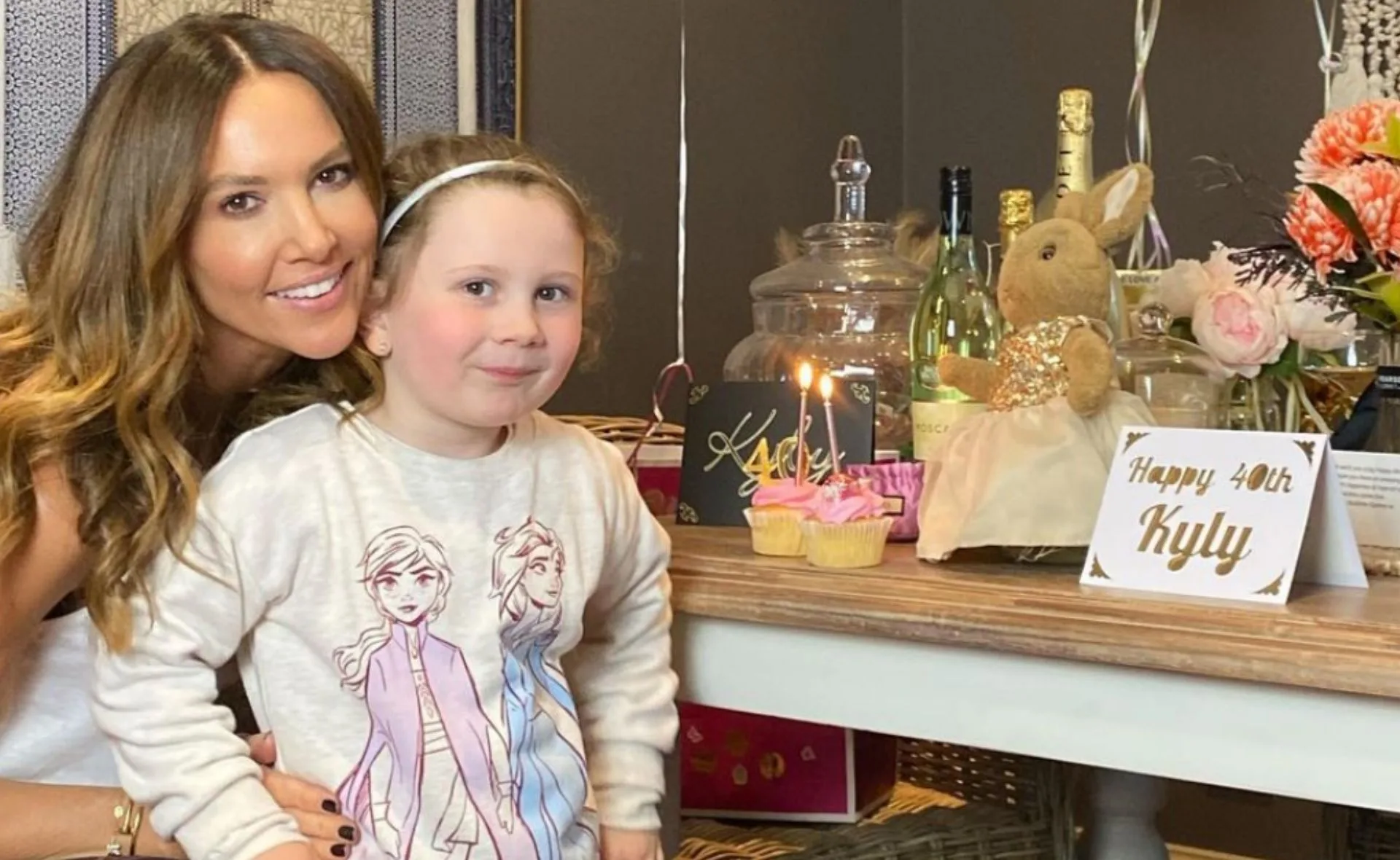 "Two people smiling at a table with birthday decorations, a 'Happy 40th Kyly' sign, and a cake with candles."