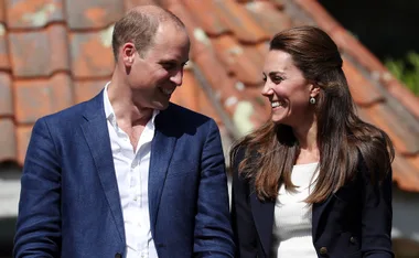 A man and woman smiling at each other outdoors, both wearing dark blazers with a tiled roof in the background.