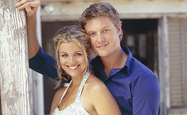 Two people smiling, one leaning against a rustic wooden post, in a setting reminiscent of "McLeod's Daughters."