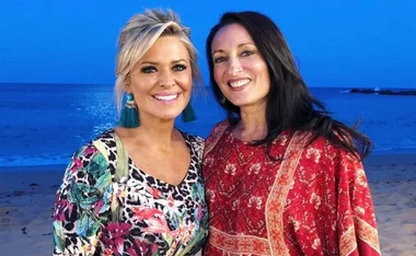 Two women smiling on a beach with a deep blue ocean and sky backdrop.