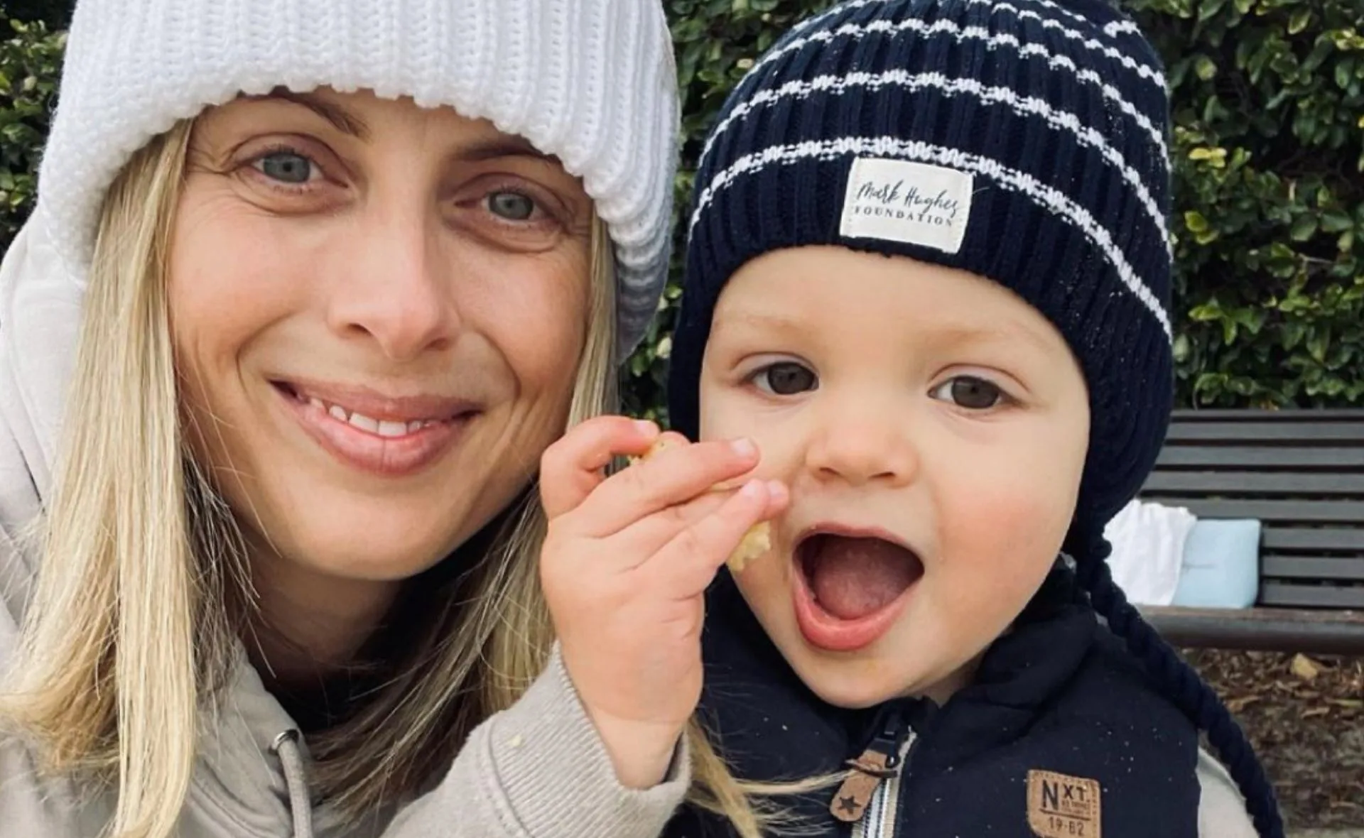 A woman and a baby both wearing knit hats smile outdoors, with the baby holding food.
