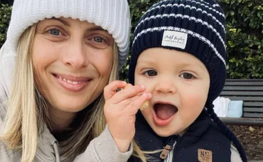 A woman and a baby both wearing knit hats smile outdoors, with the baby holding food.