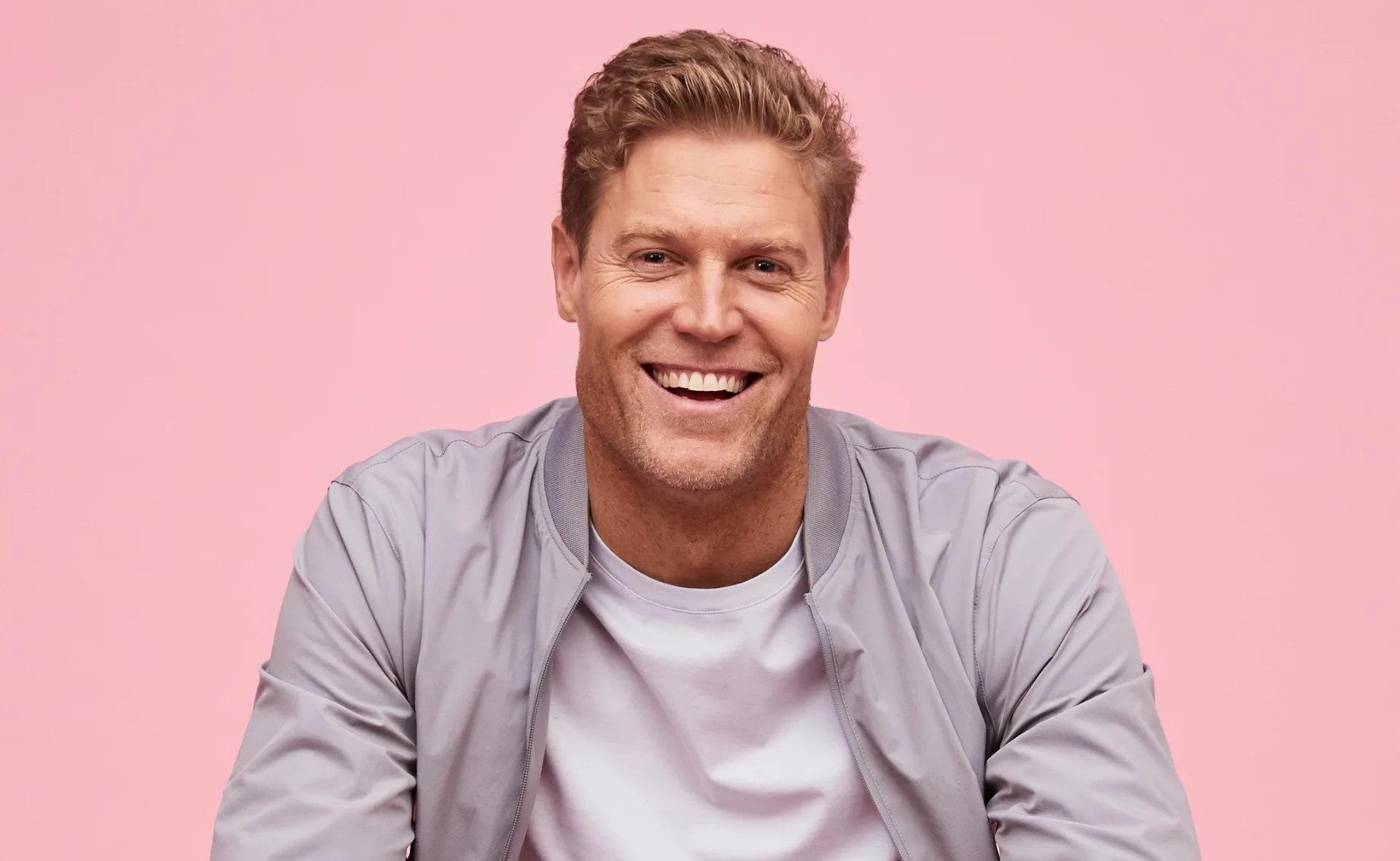 Smiling man in a light gray jacket and shirt against a pink background.