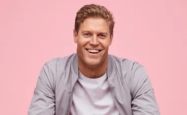 Smiling man in a light gray jacket and shirt against a pink background.