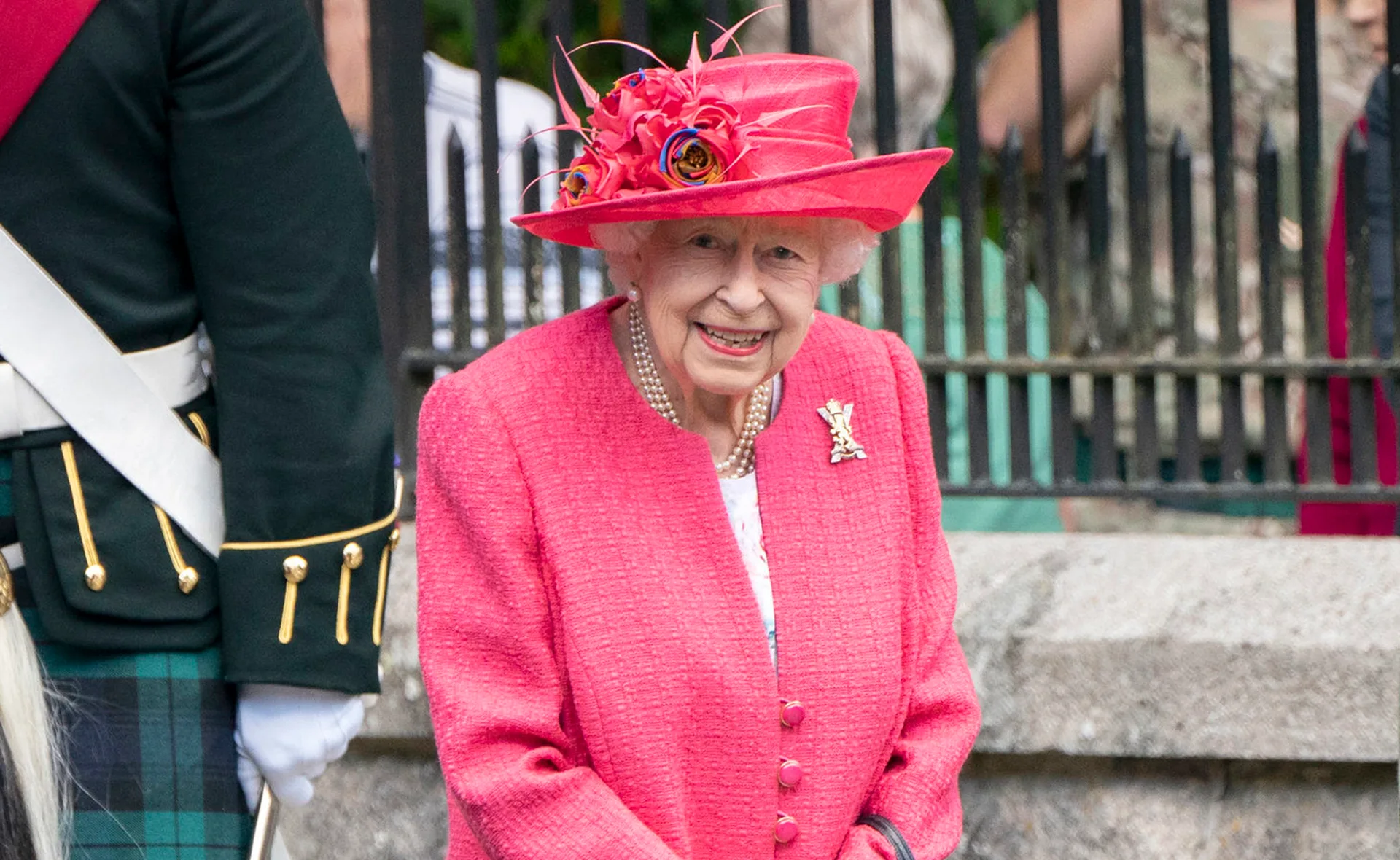 Elderly woman in a pink suit and hat, smiling, with pearls and a brooch, near a man in uniform.