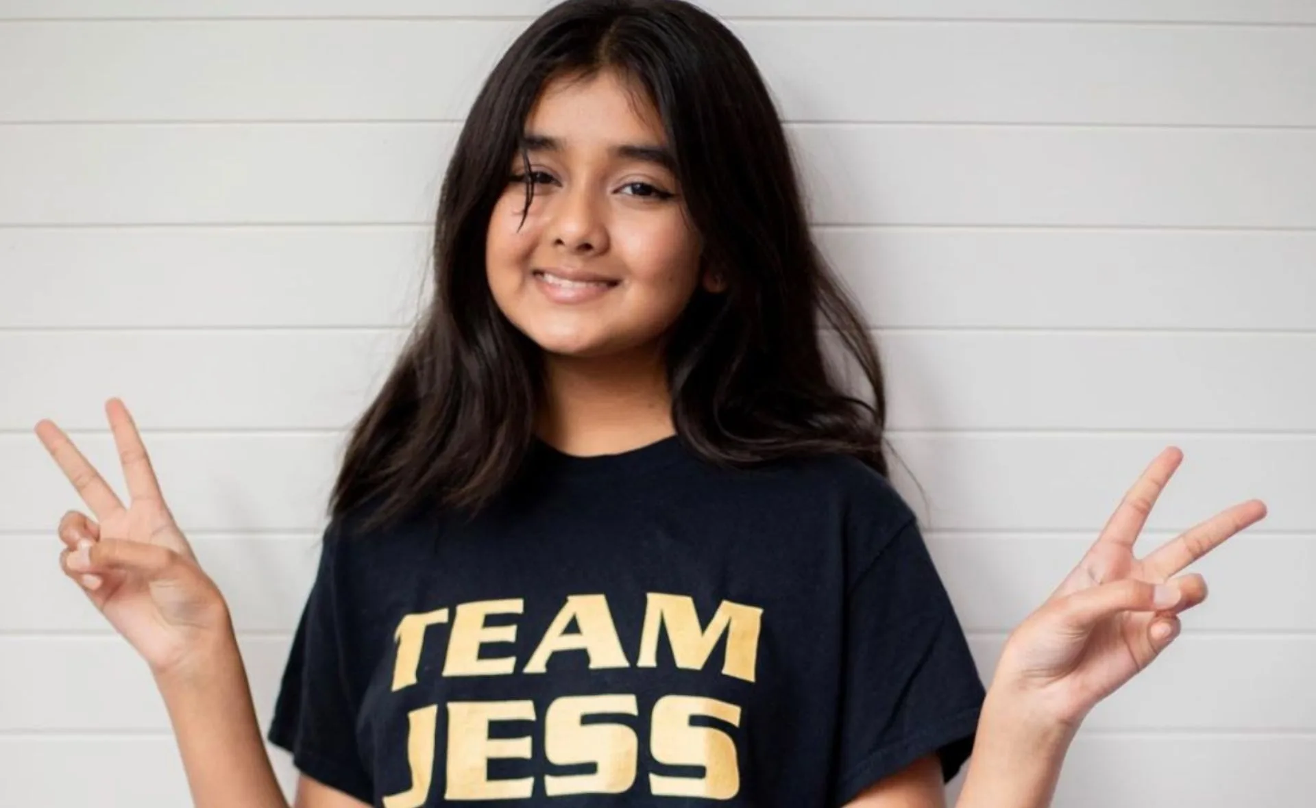 A girl smiling, wearing a "Team Jess" shirt, showing peace signs with both hands against a white wall.