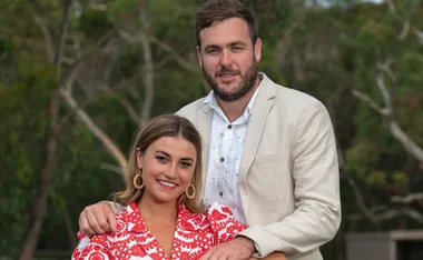 Couple posing outdoors, woman in red and white dress, man in beige jacket, smiling with trees in background.