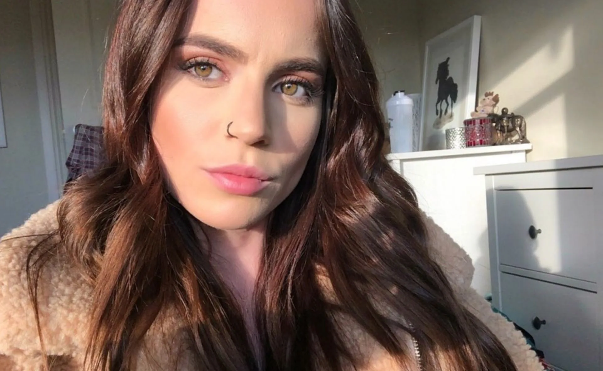 Woman with long brown hair and nose ring, seated indoors with sunlight on her face, framed picture and decor in the background.