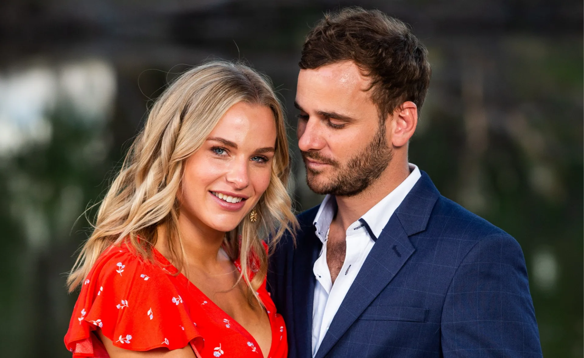 Man in blue suit gazing at smiling woman in red dress outdoors.