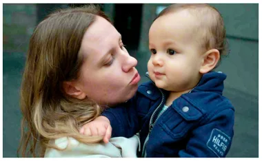 A woman holds and looks at a young child dressed in a navy blue jacket, both gazing in the same direction.