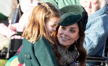 Woman in a green hat holding a young girl with braided hair, both smiling and dressed warmly at an outdoor event.