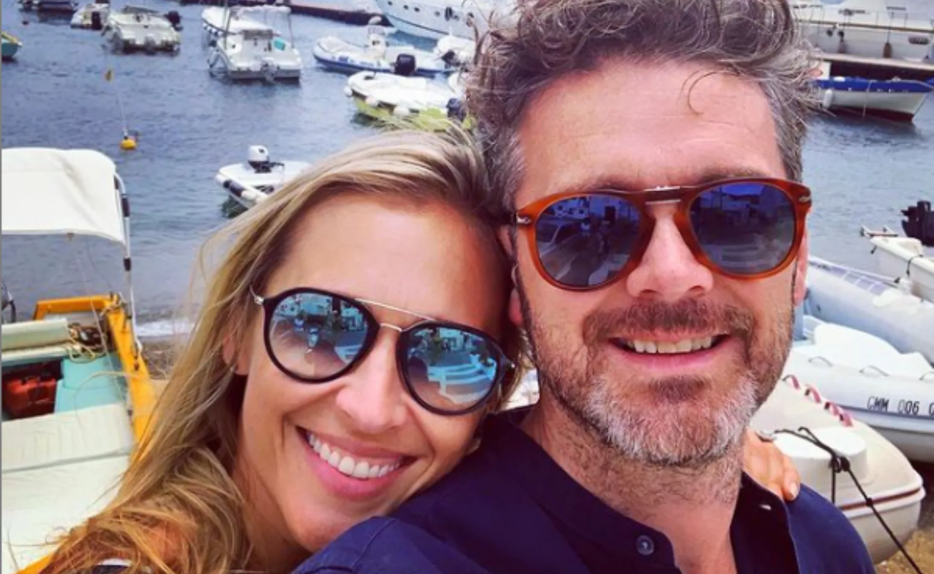Couple smiling in sunglasses with boats and water in the background.