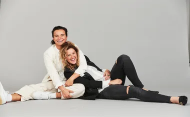 Two individuals smiling, sitting and playfully embracing each other on a gray backdrop, dressed in casual and stylish clothing.