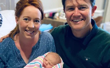 A smiling couple sit on a hospital bed, each holding a newborn wrapped in a striped blanket.