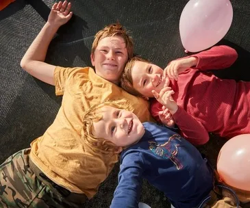Three smiling children lying on a trampoline, holding pink balloons, under a sunny sky.