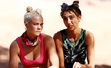 Two women sitting outdoors under sunlight with serious expressions, one in a pink top and the other in a tie-dye top.
