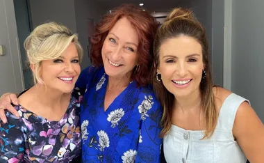 Three women smiling together, one in a floral dress, one in a blue patterned top, and one in a light-colored dress.