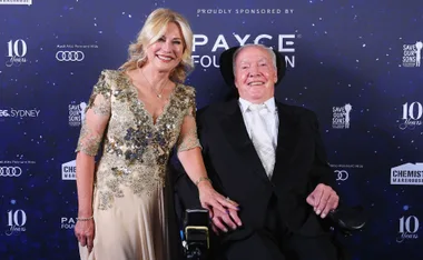 Couple smiling at a gala event; the woman in an embellished gown, the man in a suit seated in a wheelchair.