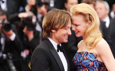A couple smiling and leaning towards each other at a formal event with photographers in the background.