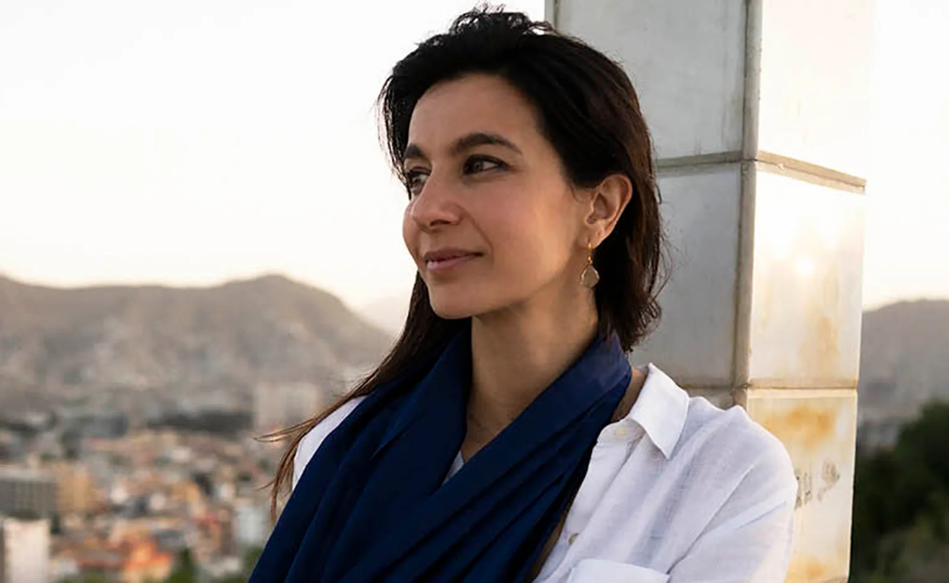 A woman in a white shirt and blue scarf gazes to the side with a cityscape and mountains in the background.