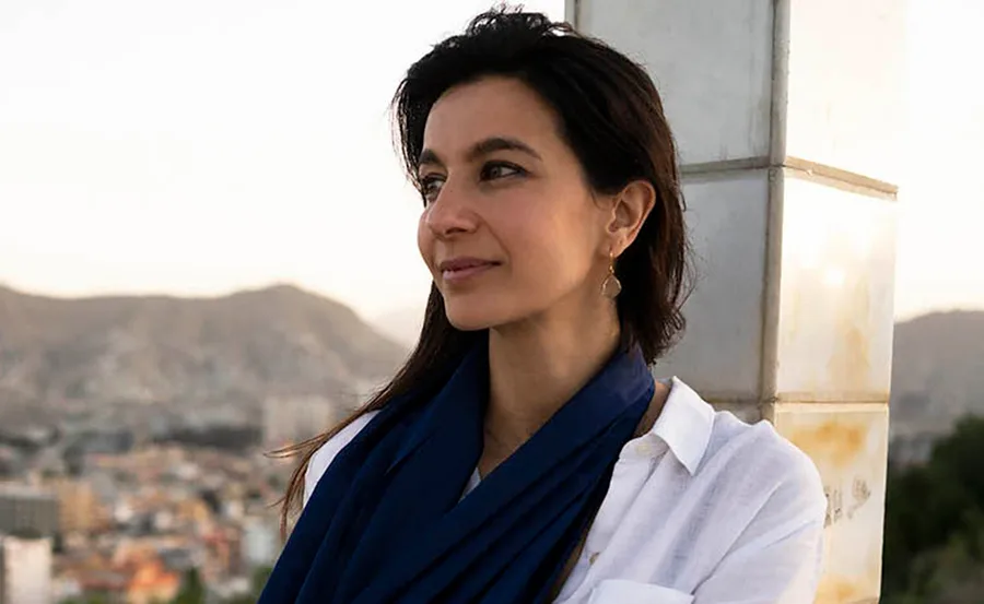 A woman in a white shirt and blue scarf gazes to the side with a cityscape and mountains in the background.