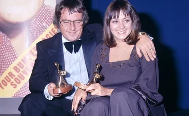 Entertainers holding awards, smiling at an event, with a promotional poster in the background.