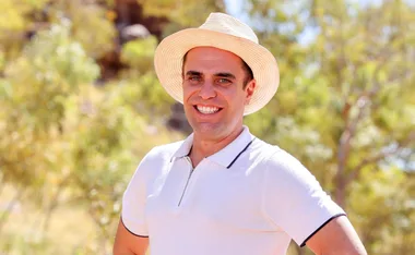 Man smiling in a white hat and shirt, outdoors with blurred greenery in background.