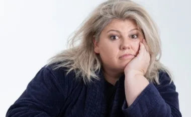 A woman with light hair looking thoughtful, wearing a dark robe, rests her head on her hand against a plain background.