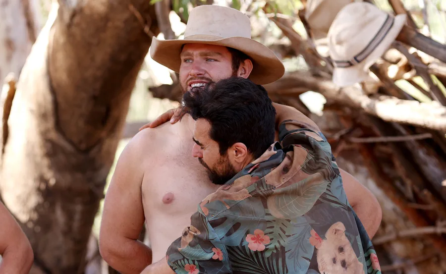 Two men embrace, one shirtless wearing a cowboy hat, in a heartfelt moment outdoors, surrounded by trees and hats.