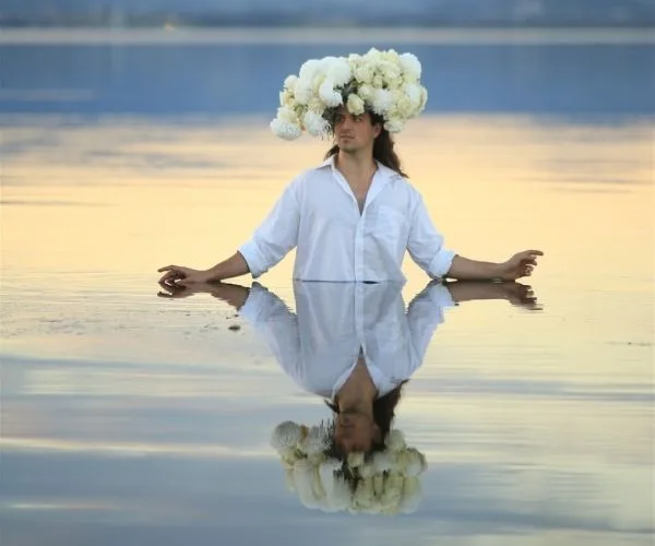 A person stands in water wearing a large floral crown against a serene sunset, with their reflection visible.