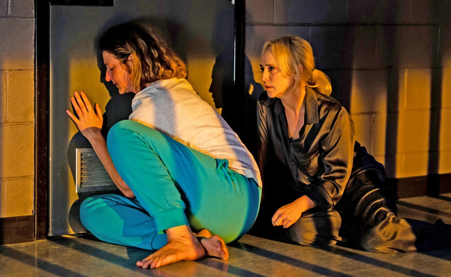 Two women sit on the floor in a dimly lit room, one listening at the door.