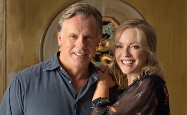 A smiling couple standing together, with the man wearing a blue shirt and the woman in a floral top.