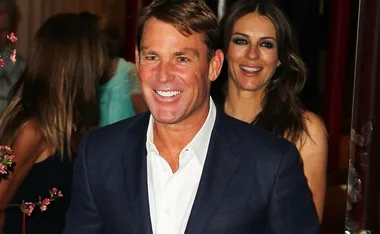 Man in a suit smiling with a woman behind him, both appear happy at an event.